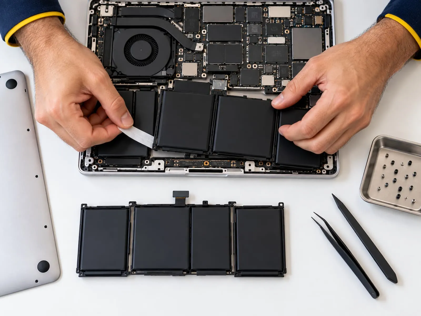 Technician removing an old MacBook battery, with the replacement battery on the desk